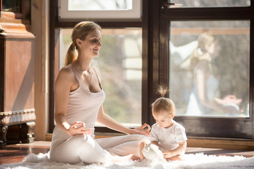 Young yogi mother in Easy Seat pose, wearing white sportswear, baby daughter playing near her,...
