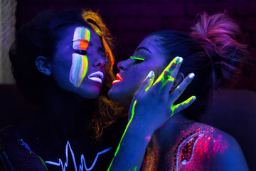 Sexy lesbian fashion models in uv neon light with fluorescent glowing Body Art make-up kissing. Low...