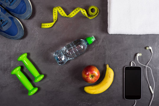 Fitness equipment background. Dumbbell, apple, banana, water bottle, towel, smartphone and headphones, sneakers on grey background. Top view