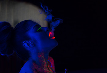 portrait of fashion model woman in uv neon light with fluorescent glowing Body Art make-up. She blows smoke from the hookah. Low key dark image. Soft focus image.