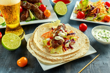 Homemade Kofta kebabs on skewers with pita, lime, vegetables, sweet chili and yogurt sauce on white plate