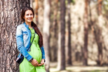 Positive fashionable pregnant woman with a smile and hands on her belly poses next to a tree in the forest in the summer