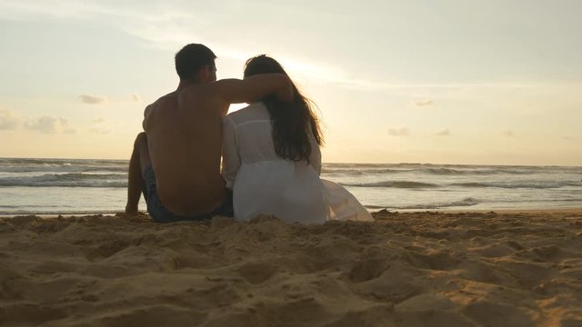 A woman and a man sits together in the sand on the sea shore, admiring the ocean and landscapes. Young romantic couple is enjoying beautiful sunset sitting on the beach and hugging. Close up