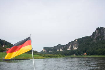 German flag at ferry through the Elbe at Kurort Rathen village and Bastei rocks at the background, Saxon Switzerland, Germany.