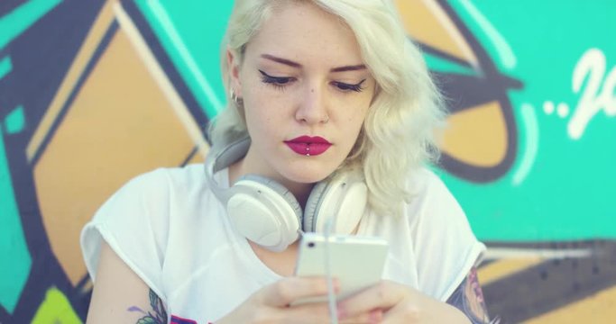 Hipster attractive young blond woman browsing her mobile for a music soundtrack as she sits in front of colorful graffiti wearing headphones round her neck