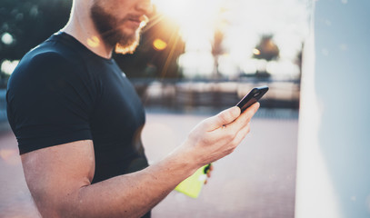 Closeup view of Muscular handsome athlete checking sport results on smartphone application and smart watch after good workout session on city park in the sunny morning.Blurred background.