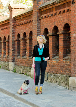 Pretty blonde girl walking with funny pug. Funny dog with tongue hanging out near brick wall on the sidewalk. Yellow shoes