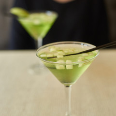 Two glasses of apple martini and apple slices on table in cafe. Young woman blurred at background