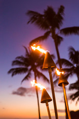 Hawaii sunset with lit tiki torches. Hawaiian icon, lights burning at dusk at beach resort or...