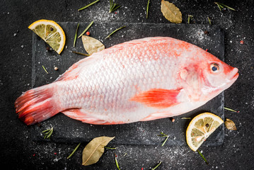 Raw fish red tilapia on a chopping board on a black stone table, with spices, lemon and herbs for cooking. Top view copy space