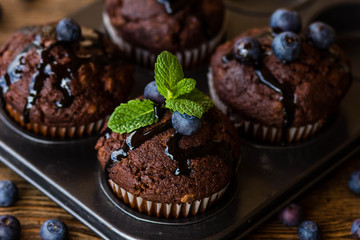 Chocolate muffins with chocolate syrup, blueberries and mint in a wooden background