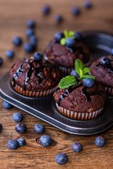 Chocolate muffins with chocolate syrup, blueberries and mint in a wooden background
