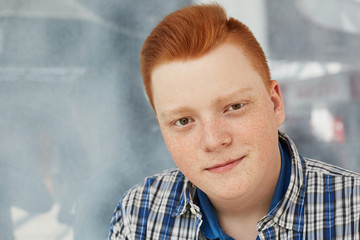 A close-up of positive guy with round face having freckles and stylish red hair dressed in fashionable checked shirt looking directly into camera.
