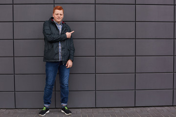 Fototapeta na wymiar A horizontal portrait of stylish redhead man dressed in checked shirt black jacket jeans and running shoes standing near black wall pointing at it with copy space for your text or advertising content.