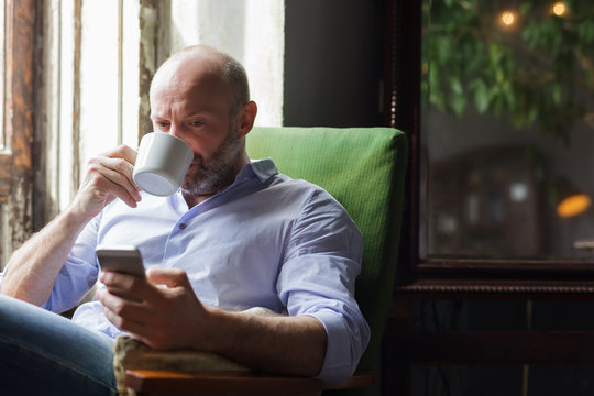 A man drinks coffee and checks the news in the phone, sitting in a chair by the window - Powered by Adobe