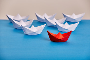 Bright red paper ship leading white ones based on blue against light background.