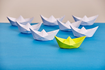 Bright yellow paper ship leading white ones based on blue against light background.
