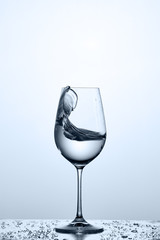 Splashing cleaner water wave in the wineglass while standing on the glass with drapes against light background.