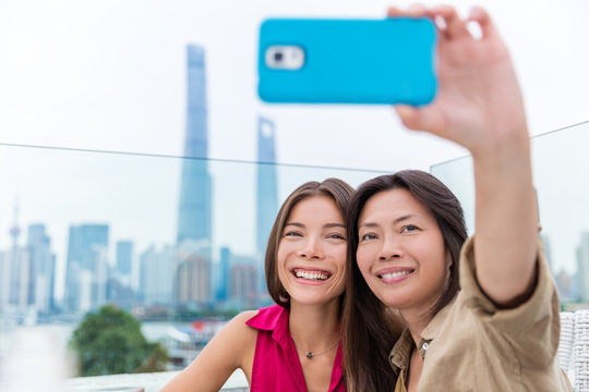 Chinese mother and daughter taking selfie together. Happy multiracial family on travel holiday in Shanghai, China. Mom holding mobile phone on outdoor cafe terrace with view of the Bund and Pudong.