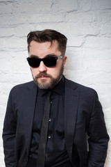 Obraz premium Portrait of a bearded serious man in sunglasses and a business black suit against a white brick wall