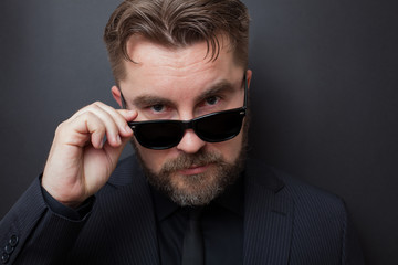 Fototapeta premium A brutal man with a beard and a stylish hairstyle in a black suit looks appallingly at the camera, dropping his sunglasses