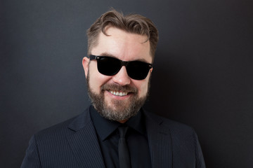 Obraz premium A young man with a beard in a stylish black business suit stands on a black background and smiles