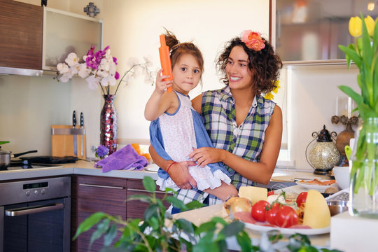 Attractive brunette female with curly hair and her cute little daughter cooking food in a home kitchen.