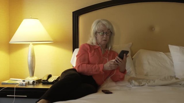 mature woman relaxing in hotel room but always plugged in via smart phone and engaged in business conversation.