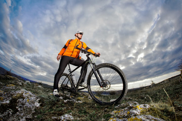 Portrait of the mountain cyclist standing with bike on the rocky hill against dramatic sky with clouds.
