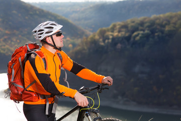 Portrait of young cyclist with a red backpack standing under river against summer landscape with...