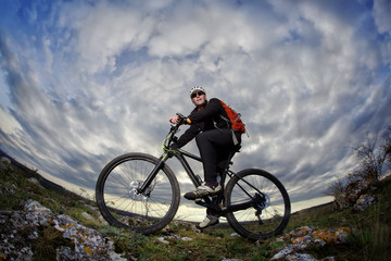 Portrait of the young cyclist standing with bike on the rocks against dramatic sky with clouds.