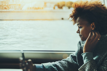 True tilt shift portrait of pensive black teenager girl using smartphone inside of ship cabin, biracial young female with cell telephone near ship window with river water outside on moody day - Powered by Adobe
