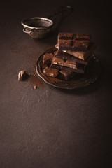 Broken chocolate pieces and cocoa powder on metal plate on brown background with copy space. Dark photo.