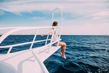 Beautiful woman in white swimsuit standing on the nose of the yacht and spreading hands apart at a sunny summer day, breeze developing hair, beautiful turquoise sea on background