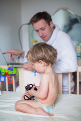 A little son sick on rashes sits sad on bed and keeps photo camera in hands, worried father sits...