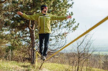 A man, aged with a beard and wearing sunglasses, balances on a slackline in the open air between two trees at sunset