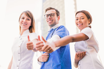 Three smiling happy doctors stand and show thumbs up outdoor