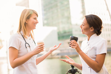 Two smiling nurses stand with cups of coffee and tell with each other during the coffee break, outdoors