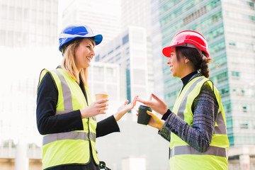 Two young smiling engineers stand together with cups of coffee in hands and discuss about something during break outdoor