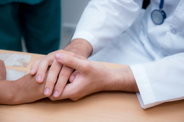 Friendly male doctor hand holding patient hand sitting at the desk for encouragement - empathy - cheering and support while medical examination.