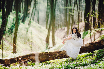 Woman in elegant white dress sits on the tree in sunny day