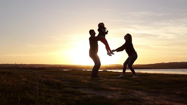 Silhouette, happy child with mother and father, family at sunset, summertime. Run, raising baby up in the air, hugs, love, playing. We are happy family.