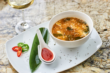 Korean cuisine. Spicy kimchi soup in white ceramic bowl served with red sauce in spoon, green leaf and paprika rings with glass of white wine on marble table.