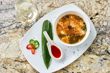 Korean cuisine. Spicy kimchi soup in white ceramic bowl served with red sauce in spoon, green leaf and paprika rings with glass of white wine on marble table.