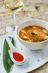 Korean cuisine. Spicy kimchi soup in white ceramic bowl served with red sauce in spoon, green leaf and paprika rings with glass of white wine on marble table.