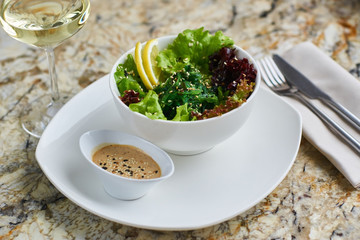 Seeweed salad in white ceramic bowl served with lemon slices, sauce, glass of white wine, fork and knife on marble table