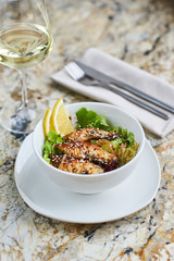 Asian cuisine. Warm salad with eel fish and rice served in white ceramic bowl with knife, fork and glass of white wine on marble background