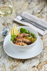 Asian cuisine. Warm salad with eel fish and rice served in white ceramic bowl with knife, fork and glass of white wine on marble background