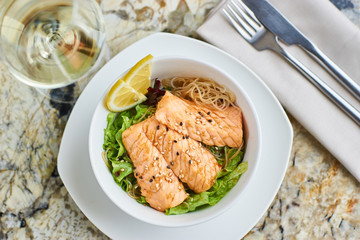 Asian cuisine. Warm salad with salmon fish and rice served in white ceramic bowl with knife, fork and glass of white wine on marble background. Flat lay or top view.