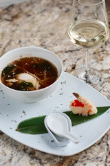 Japanese cuisine. Noodle soup udon in white ceramic bowl served with spoon, glass of white wine and king prawn on marble table